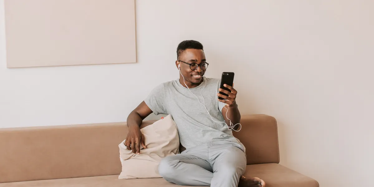 A man in a grey t-shirt with wired earphones sits on a tan sofa, smiling at the phone in his hand.
